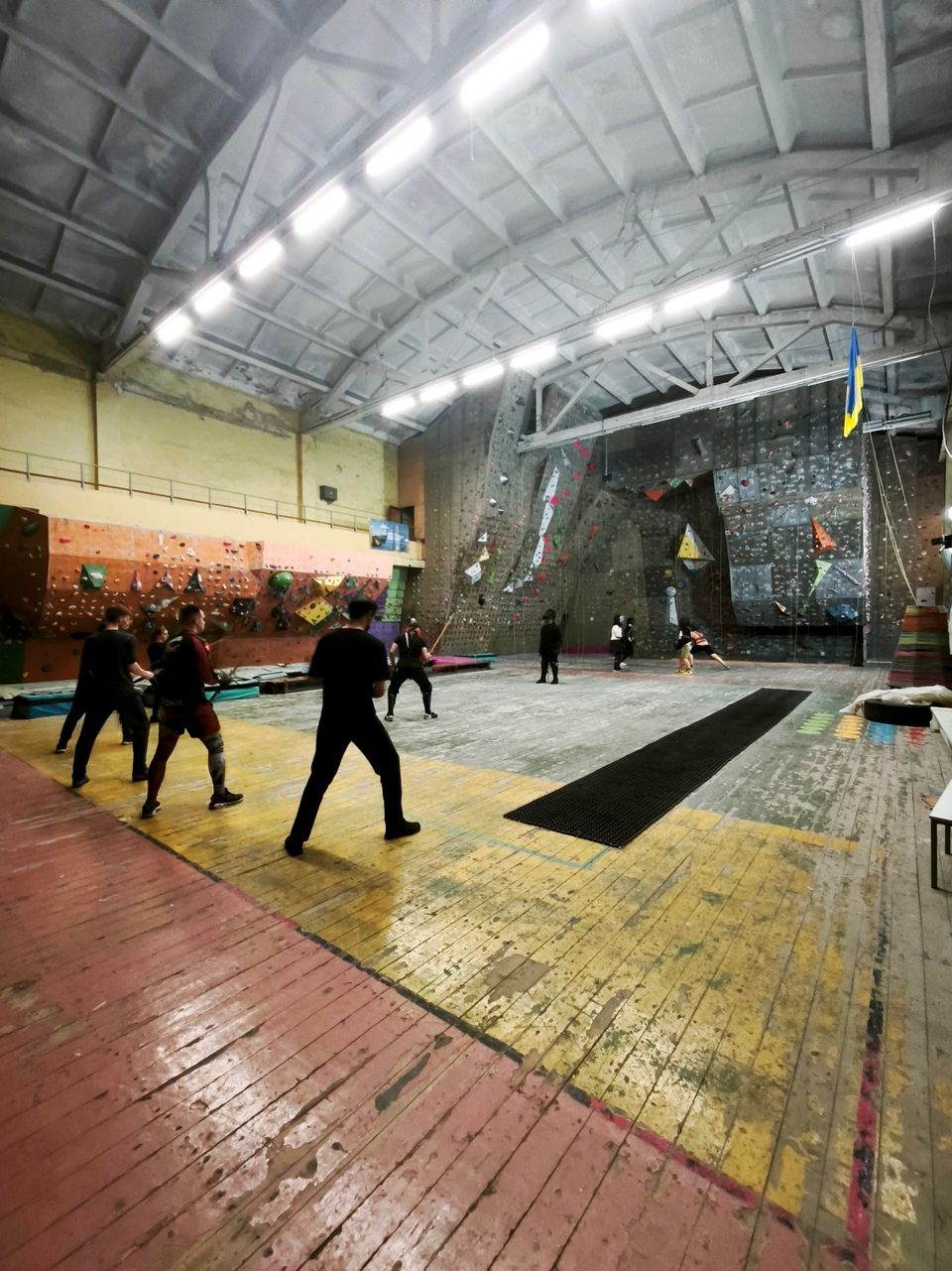 Atira Ruthenia group at a historical fencing training session in the large NAU climbing hall in Kyiv: members in black practise drills, with masked fencers and climbing walls behind them