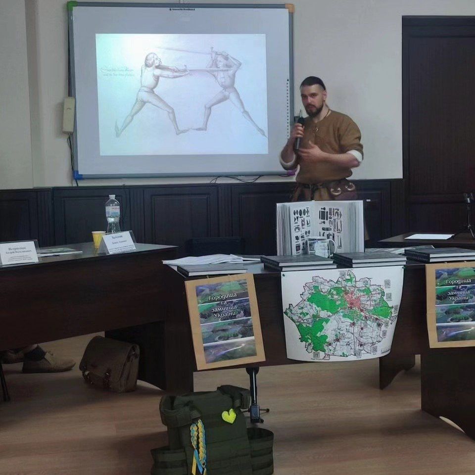 A club member in medieval clothing presenting Atira Ruthenia at the Drevlian Towns conference in Korosten, with a historical fencing illustration on screen