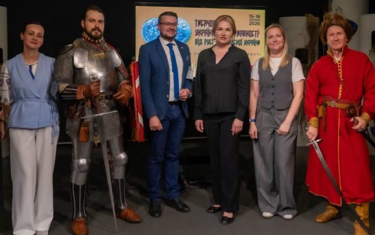 Six people stand in a row in front of the festival banner: a man in full plate armour holding a sword, a man in a long red kaftan with a sabre, and four guests in modern dress.